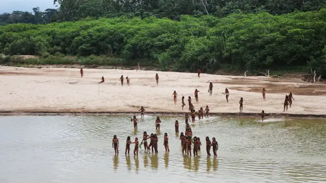 Un grupo de personas de pie, en aguas poco profundas cerca de la orilla de un río arenoso. Hay más personas dispersas sobre la arena, con un denso bosque verde al fondo.