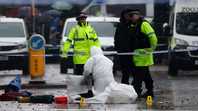 Dos agentes de policía y otra persona en pie en una calle. Otra persona, vestida de blanco, arrodillada frente a ellos, trabaja en la escena del crimen en que se produjo el ataque a la sinagoga de Manchester.