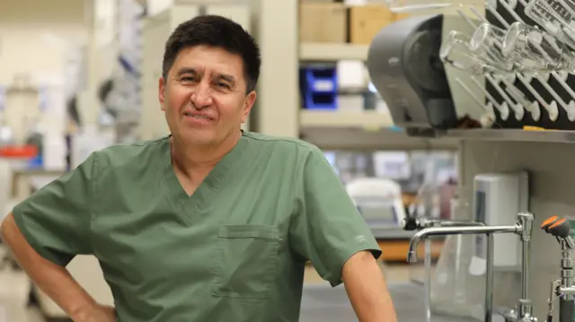 Hombre con uniforme verde oliva en un laboratorio con grifos y equipo científico en el fondo, sonriendo a la cámara.
