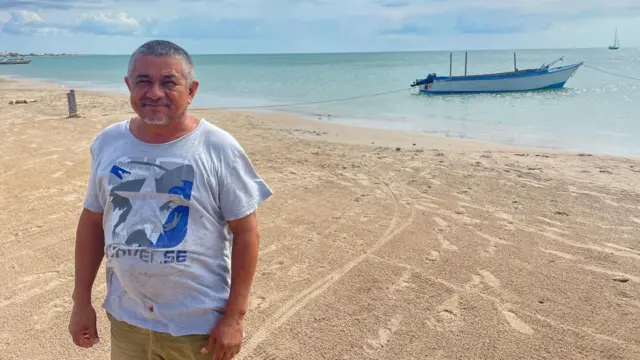 El pescador wayuu Aaron Laguna de pie en una playa con su bote en el mar detrás de él.