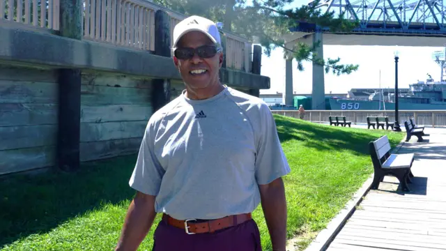 Tom Teixeira, con una camiseta gris y pantalones cortos, camina junto al río en Fall River, con el puente Braga de fondo.