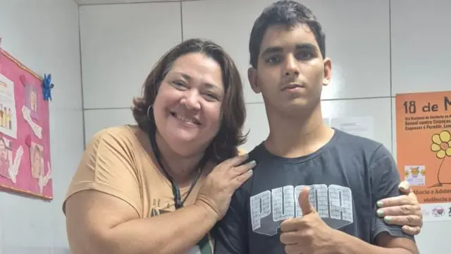 Verónica Oliveira y Gerson de Melo Machado posan para una foto juntos; ella sonríe mientras le da un abrazo, mientras él muestra su pulgar hacia arriba