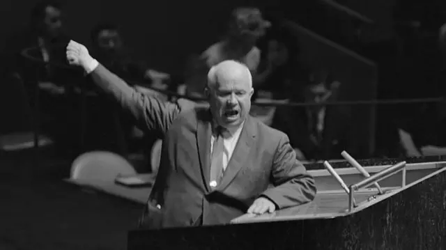 Nikita Khrushchev se dirige a la Asamblea General de las Naciones Unidas desde el podio de oradores. Mientras gesticula tiene el rostro serio. Apoya su brazo izquierdo sobre la mesa, mientras levanta en alto el puño de su brazo derecho.