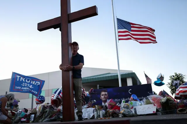 Una bandera estadounidense ondea a media asta mientras un hombre lleva una gran cruz cerca de un monumento improvisado para el fundador de Turning Point USA, Charlie Kirk, afuera de la sede de Turning Point USA en Phoenix, Arizona, el 12 de septiembre de 2025.