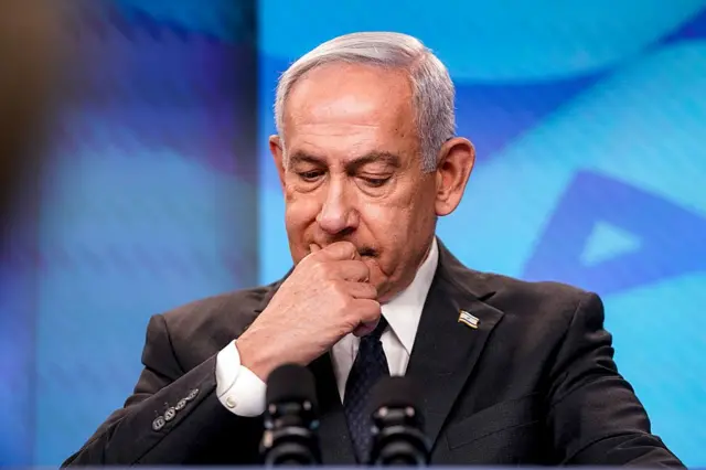 El primer ministro de Israel, Benjamin Netanyahu, vestido con traje oscuro, está sentado ante unos micrófonos, con una mano cubriendo la boca en un gesto serio y pensativo.