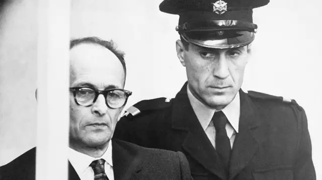 Adolf Eichmann tras una barra con un agente de seguridad uniformado detrás de él.