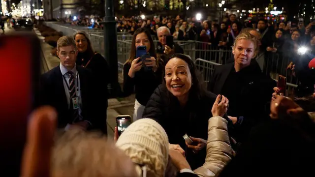 María Corina Machado sonríe mientras saluda a un grupo de personas que le esperaba frente a su hotel en Oslo, Noruega. Viste un abrigo negro. 