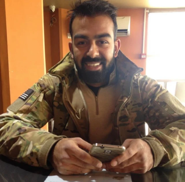Safi, vestido con uniforme militar, sonriendo a la cámara mientras sostiene un teléfono.