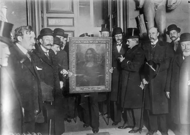 Imagen en blanco y negro de 1914 de un grupo de hombres vestidos con elegantes abrigos, trajes, corbatas y sombreros negros. En el centro, una imagen original enmarcada de la Mona Lisa
