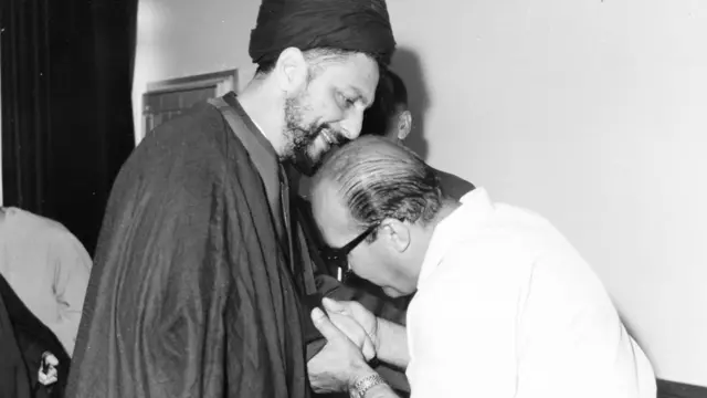 Sadr, sonriente, se inclina hacia un hombre con anteojos y vestido de blanco que sostiene las manos de Sadr para besarlas
