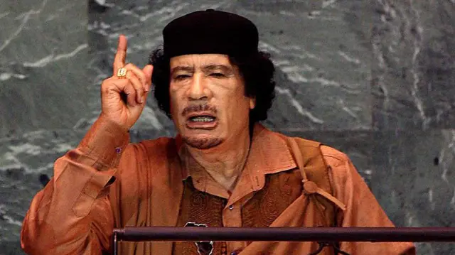 Muamar Gaddafi luciendo un sombrerito negro y vistiendo un traje color ocre, habla desde el podio de la ONU. Tiene la mano derecha levantada con el dedo índice apuntando hacia arriba y en el dedo anular lleva un anillo dorado.