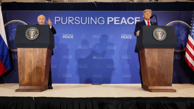 Putin habla desde un podio y señala con la mano izquierda mientras Trump está de pie en otro podio a la izquierda de Putin y mira hacia adelante, en Anchorage, Alaska, el 15 de agosto de 2025. Un letrero detrás de ellos dice 