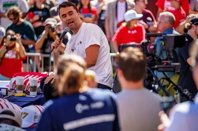 Charlie Kirk con una camiseta blanca y un micrófono en la mano se dirige a una multitud en un mitin.