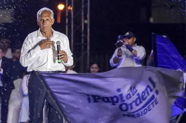 Nasry Asfura en un escenario durante la campaña electoral. Frente a él se ve una bandera en la que dice: 
