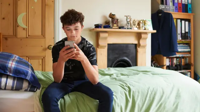 Un adolescente viendo un celular sentado sobre una cama.