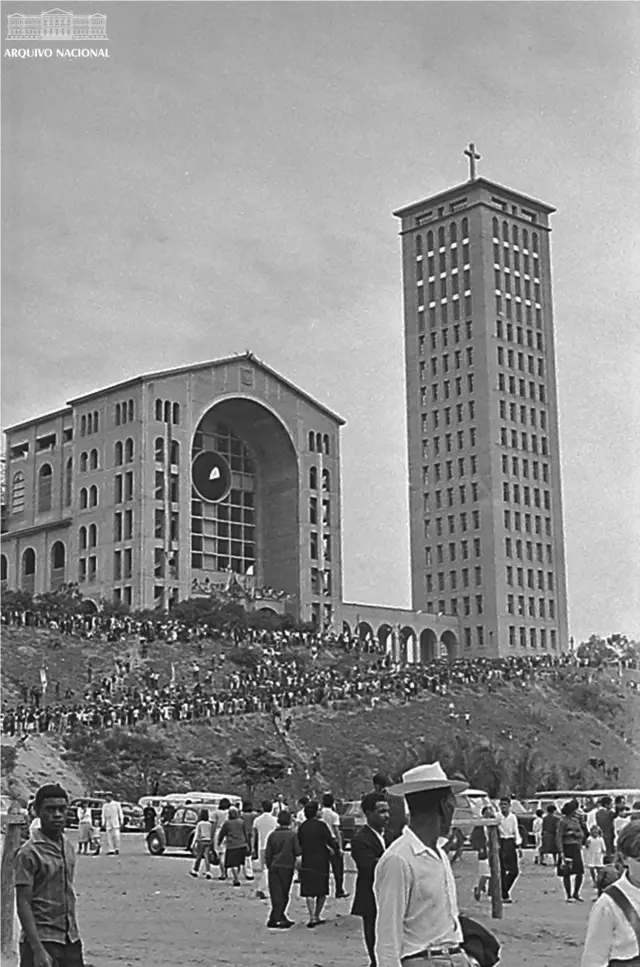 Fotografía en blanco y negro de personas caminando. Al fondo se ven edificios y una multitud.