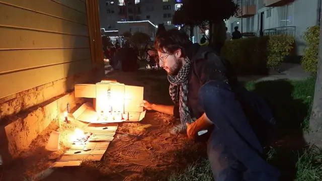 Manuel Tique, tumbado sobre el césped durante la noche frente a lo que parece ser un pequeño memorial con flores y velas.