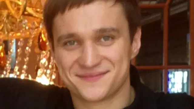 Un hombre con el pelo castaño muy corto sonríe frente a una gran lámpara de araña.