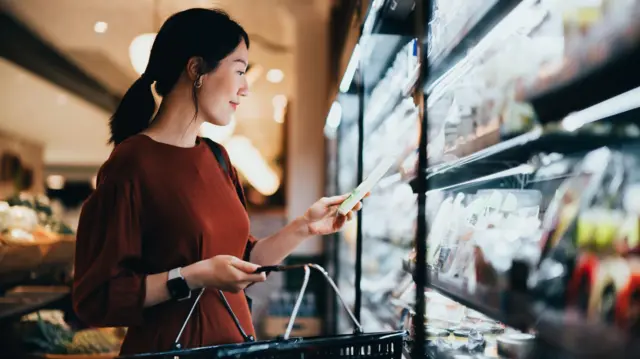 Una mujer observa un producto que lleva en la sección refrigerada de un supermercado. Lleva un vestido marrón y sostiene una cesta en una mano.