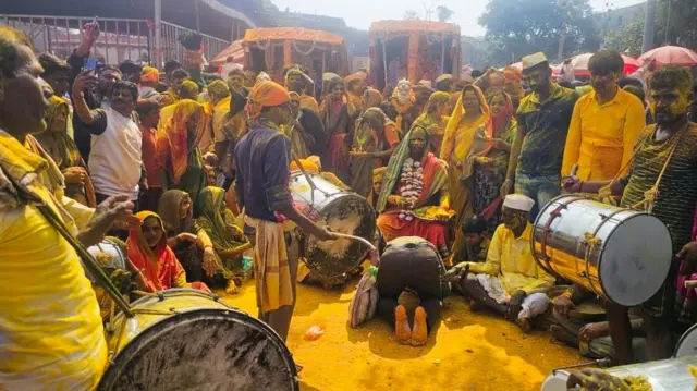 Un grupo de hombres y mujeres están sentados en semicírculo alrededor de una anciana sentada en una silla. Todos tienen la cara cubierta de polvo amarillo. Tres hombres en primer plano tocan tambores.