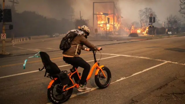 Un hombre huye del incendio en Pacific Palisades en bicicleta y pasa frente a un edificio en llamas en Sunset Boulevard en medio de fuertes ráfagas de viento. Los Ángeles, California, Estados Unidos, 8 de enero de 2025. (Foto: Apu Gomes/Getty Images)