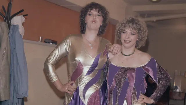 Dos concursantes del Intervision de 1977 posan para la cámara entre bastidores. La más alta, con cabello castaño, frunce los labios. La más baja, con cabello rubio, sonríe. Las dos llevan trajes a juego de color dorado y morado. Al fondo se ve un perchero con abrigos.