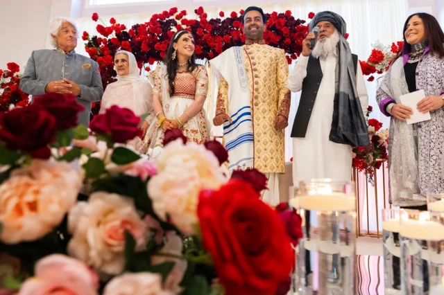 Sammi y Safi de pie en el escenario con sus respectivos padres durante la celebración de su boda. Hay un fondo de flores rojas y un primer plano borroso de flores de colores.