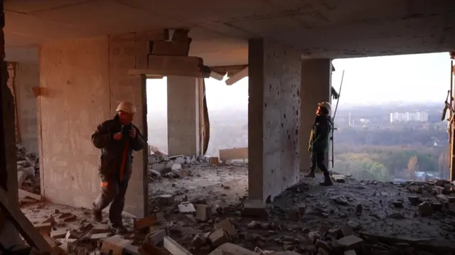 Dos bomberos con trajes de protección recorren un edificio destruido a gran altura. Las ventanas han estallado y, a través de los huecos en la pared, se ven edificios y árboles.