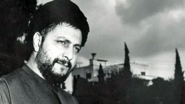 Foto en blanco y negro de Musa al-Sadr. Tiene barba y turbante negro y se ve pensativo. En el fondo se ven árboles y edificios  