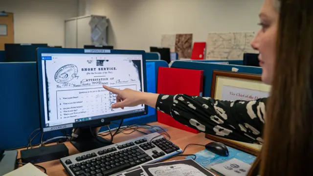 Nicola Nash sentada en un escritorio rodeada de documentos y libros. Lleva una blusa blanca y negra y tiene el pelo largo y castaño. Señala un ordenador de sobremesa con un documento histórico en la pantalla, el historial de servicio del soldado Gordon McPherson.