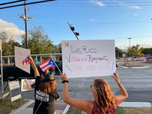 personas con carteles y banderas de puerto rico. Las cartulinas dicen 