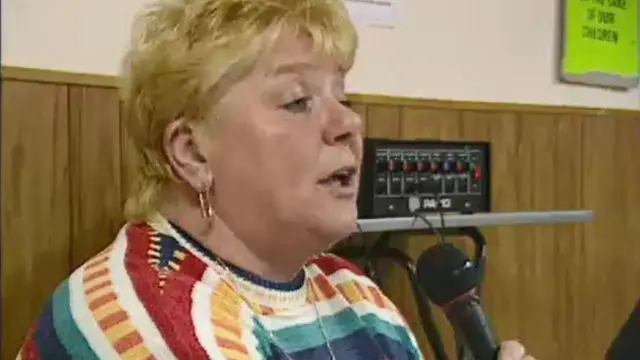 Una captura de pantalla de Haney dirigiéndose a un público. Sostiene un micrófono. Tiene el pelo corto y rubio y un gran aro dorado. Lleva un jersey multicolor.