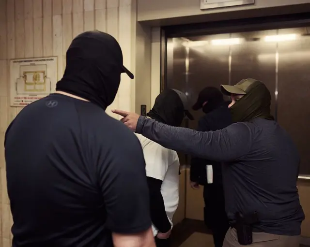 Cuatro agentes migratorios con las caras tapadas entran en un ascensor. 