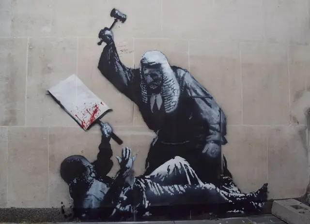 La última obra del artista callejero Banksy aparece en un muro de los Reales Tribunales de Justicia, representando a un juez golpeando a un manifestante con un mazo.