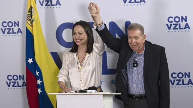 Maria Corina Machado y Edmundo González levantan sus brazos agarrados de la mano. En el fondo, una bandera de Venezuela y un logo que dice 