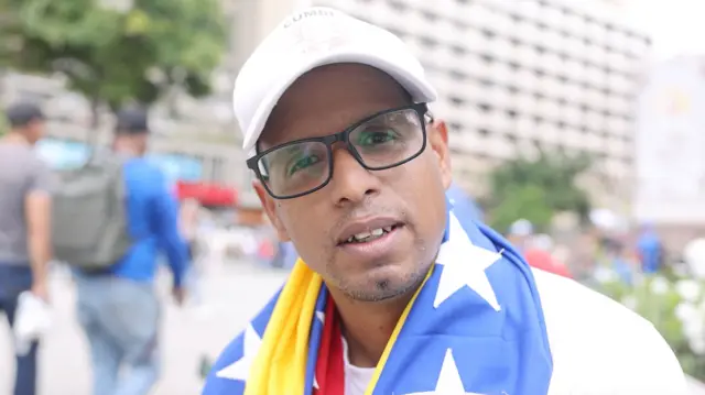 Un hombre con gorra blanca y la bandera de Venezuela en los hombros mira a cámara.