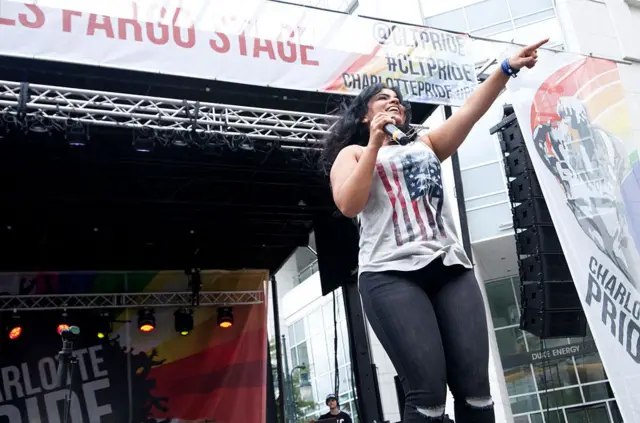 La cantante Vassy señala a la audiencia desde un escenario. Lleva una camisilla blanca con una bandera estadounidense estampada. Detrás de ella hay un escenario con una pancarta arcoíris que dice 
