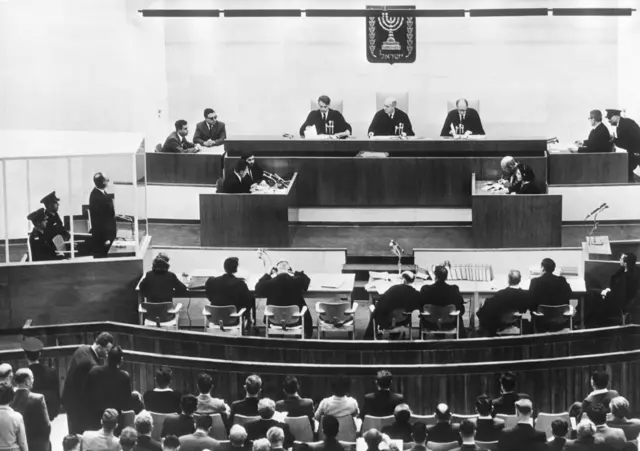 Un plano general de la sala del juicio en la que se ve Eichmann en su cubículo, lo jueces y otros miembros