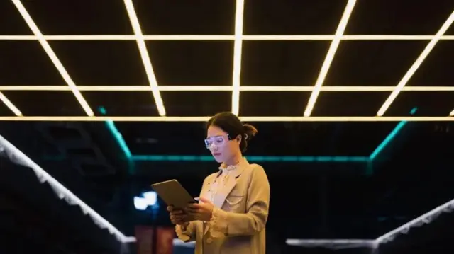 Una mujer con gafas transparentes y una tableta en la mano, en un salón oscuro con varias lámparas de luz fluorescente en el techo, las cuales le dan una sensación futurista a la foto.