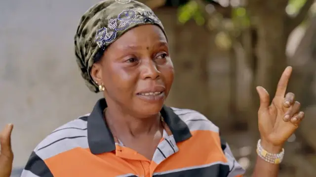 Sally Kalokoh, con un pañuelo en la cabeza y una camisa a rayas blancas, negras y naranjas, y un cuello negro, gesticula con las manos. En uno de sus brazos lleva un brazalete de cuentas que parecen perlas.