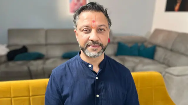 Sanjiv Patel vestido con una camisa azul y sentado en un sofá amarillo, con un sofá gris con cojines al fondo.