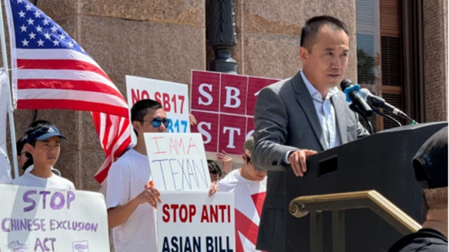 Jason Yuan da un discurso en un atril, con manifestantes detrás de él sosteniendo carteles.