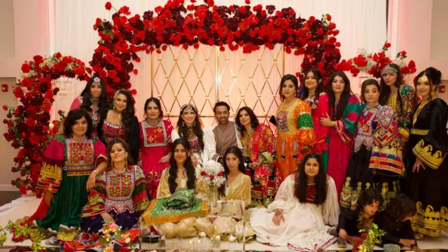 Sammi y Safi posan en su boda con sus invitados con un fondo de flores rojas en un salón de banquetes. Sus invitados visten trajes afganos.