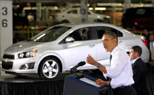 Barack Obama en camisa con las mangas dobladas, levanta el pulgar mientras da un discurso desde un podio en una fábrica automotriz con un auto color plateado en el fondo