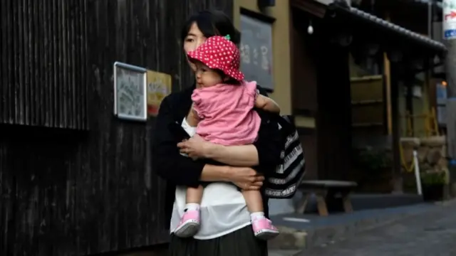 Una mujer camina por las calles de una ciudad en Japón, cargando a una pequeña bebé quien lleva un vestido enterizo rosa y un sombrero de fresa.