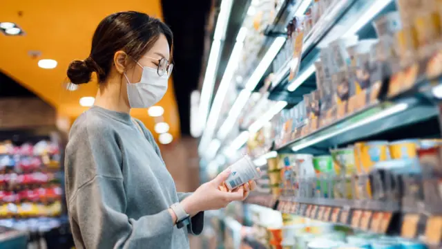 Una mujer joven se encuentra junto a la sección refrigerada de un supermercado, sosteniendo un frasco de yogur orgánico fresco y saludable, leyendo la etiqueta nutricional y comprobando los ingredientes en la parte posterior.