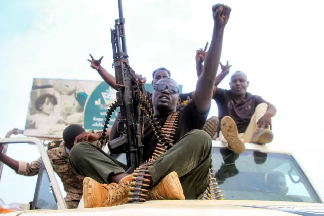 Varios hombres, el primero armado con una ametralladora y con una cinta de balas sentados en el capó de una camioneta. 