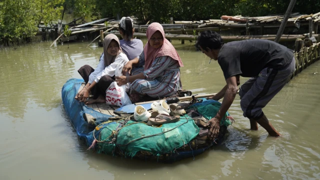 Un hombre empuja una pequeña balsa casera sobre el agua con Ningsih, su familia y una pila de zapatos a bordo.