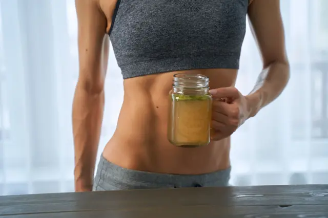 Una mujer muy marcada, con ropa de ejercicio, sostiene una taza transparente con caldo de costilla.