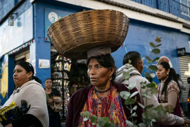 Una mujer indígena guatemalteca, vestida en un traje colorido típico y con un cesto en la cabeza, camina entre otras personas por un mercado en San Juan Sacatepequez, Guatemala